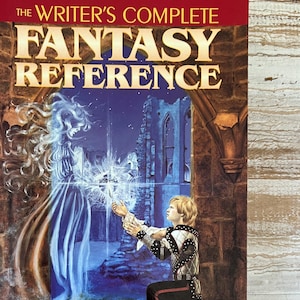 May include: A red hardcover book titled "The Writer's Complete Fantasy Reference" with a fantasy illustration of a woman in a white gown and a man in armor. The book is an "Indispensable Compendium of Myth and Magic" from the editors of Writer's Digest Books. The book has an introduction by Terry Brooks.
