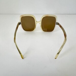 K&ouml;nnte beinhalten: Quadratische Sonnenbrille mit hellbeigem Rahmen und braunen Gl&auml;sern. Die B&uuml;gel sind hellbeige mit einer leichten Kr&uuml;mmung. Die Sonnenbrille ist vor einem wei&szlig;en Hintergrund platziert.