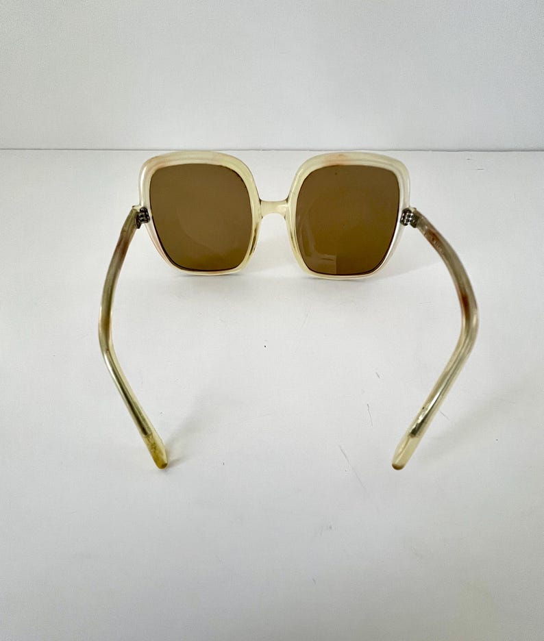 K&ouml;nnte beinhalten: Quadratische Sonnenbrille mit hellbeigem Rahmen und braunen Gl&auml;sern. Die B&uuml;gel sind hellbeige mit einer leichten Kr&uuml;mmung. Die Sonnenbrille ist vor einem wei&szlig;en Hintergrund platziert.