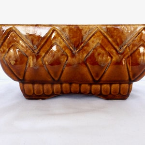 May include: A brown ceramic planter with a geometric diamond pattern. The planter has a rectangular shape with a slightly rounded bottom.