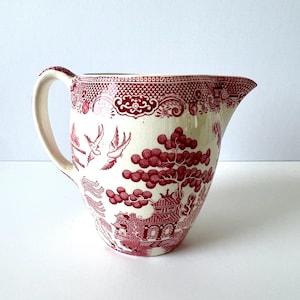 May include: Cream-colored pitcher with a red Willow pattern design. The pitcher features a handle and a spout. The design includes birds, trees, and a pagoda. The rim has a decorative border. A classic piece for serving beverages.