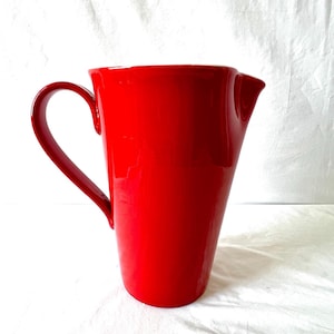 May include: A tall, glossy red ceramic pitcher with a curved handle and a pouring spout. The pitcher has a smooth, tapered design and is set against a white background. The handle is a deep red, matching the body of the pitcher.