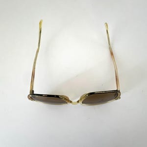 K&ouml;nnte beinhalten: Vintage Sonnenbrille mit hellbraunem Rahmen und dunklen Gl&auml;sern. Die B&uuml;gel der Sonnenbrille sind von transluzenter hellbrauner Farbe. Die Sonnenbrille ist vor einem wei&szlig;en Hintergrund platziert.