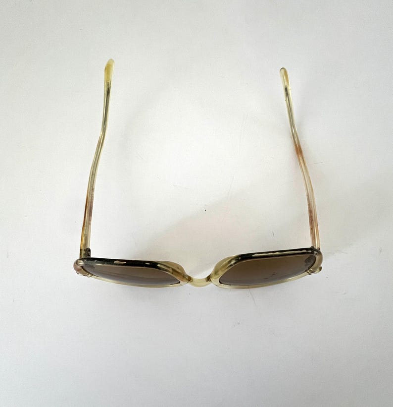 K&ouml;nnte beinhalten: Vintage Sonnenbrille mit hellbraunem Rahmen und dunklen Gl&auml;sern. Die B&uuml;gel der Sonnenbrille sind von transluzenter hellbrauner Farbe. Die Sonnenbrille ist vor einem wei&szlig;en Hintergrund platziert.