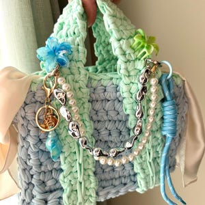 Könnte beinhalten: Handgefertigte Häkeltasche in Blau- und Mintgrüntönen, verziert mit dekorativen Anhängern und einer Perlen- und Silberkette. Die Tasche hat eine blaue Blume und ein blaues Kordeldetail, mit einem cremefarbenen Satinband-Akzent.