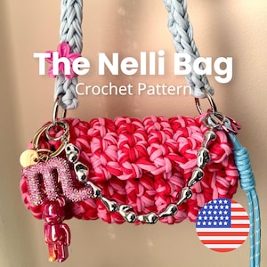 Könnte beinhalten: Eine Häkeltasche mit einer Textur aus rosa und rot meliertem Garn und einem hellblauen geflochtenen Riemen. Die Tasche ist mit einer silbernen Metallkette, einem rosa Strass-Buchstaben M und einer kleinen rosa Bärenfigur verziert. Text The Nelli Bag.