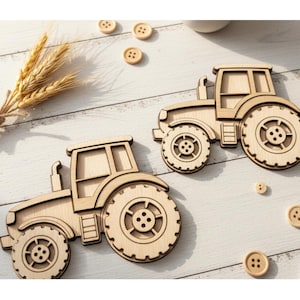 Puede incluir: Dos recortes de tractor de madera clara con diseños detallados de ruedas, colocados sobre una superficie de madera blanca. Varios botones de madera pequeños y un manojo de trigo completan la escena.