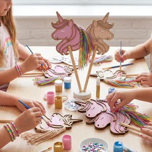 Op de afbeelding: Een knutselset met houten eenhoornhoofden op stokjes, meerkleurig garen voor manen, kleine verfpotjes in goud, roze en blauw, penselen en plaksteentjes. Een houten bordje bevat de tekst Rainbow Mane Craft Kit.