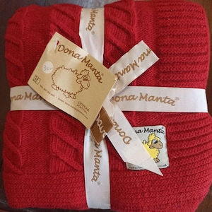 Pode incluir: Um cobertor vermelho tricotado dobrado com um design de malha de cabo, amarrado com uma fita branca impressa com "Dona Manta". Uma etiqueta com um logotipo de ovelha e o texto "Dona Manta" está anexada.