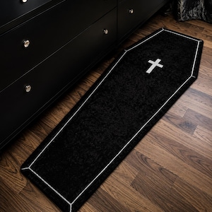 May include: Black coffin-shaped rug with a white cross and trim. The rug is placed on a wooden floor next to a black dresser. The rug's design is a unique decorative accent.