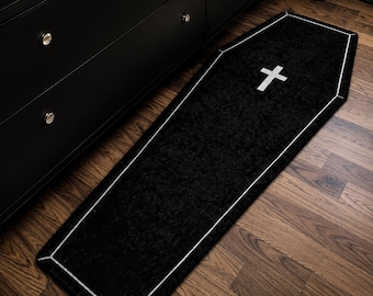 May include: Black coffin-shaped rug with a white cross and trim. The rug is placed on a wooden floor next to a black dresser. The rug's design is a unique decorative accent.