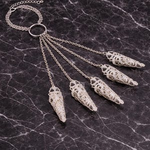 May include: Silver-toned necklace featuring a circular pendant connected to multiple chains. Each chain ends with a decorative, openwork cone-shaped element. The jewelry is displayed against a dark, marbled surface.