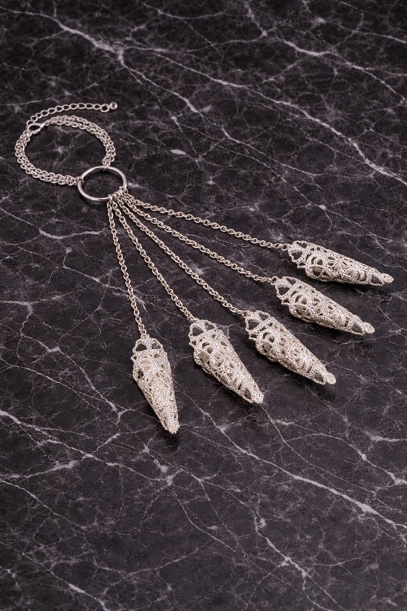 May include: Silver-toned necklace featuring a circular pendant connected to multiple chains. Each chain ends with a decorative, openwork cone-shaped element. The jewelry is displayed against a dark, marbled surface.