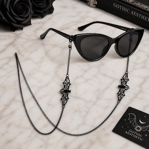 Puede incluir: Gafas de sol negras de ojo de gato con una cadena decorativa con dijes en forma de murciélago. La cadena está unida a las monturas y cuelga por debajo. Se ve una pequeña tarjeta con el texto "GOTHIC AESTHETIC".