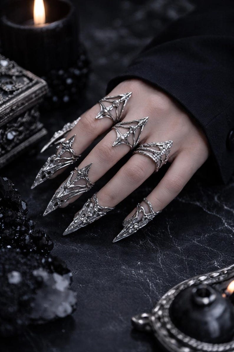 May include: A set of silver-toned metal rings designed to cover the fingers like armor. Each piece features intricate, pointed, claw-like extensions that taper to a sharp tip, with openwork detailing and textured surfaces across the finger joints.