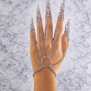 Könnte beinhalten: Silberfarbener Fingerschmuck mit aufwendigen Filigranmustern. Der Handschmuck hat Kettenglieder, die die Fingerkappen mit einem Ring und einem Armband verbinden.