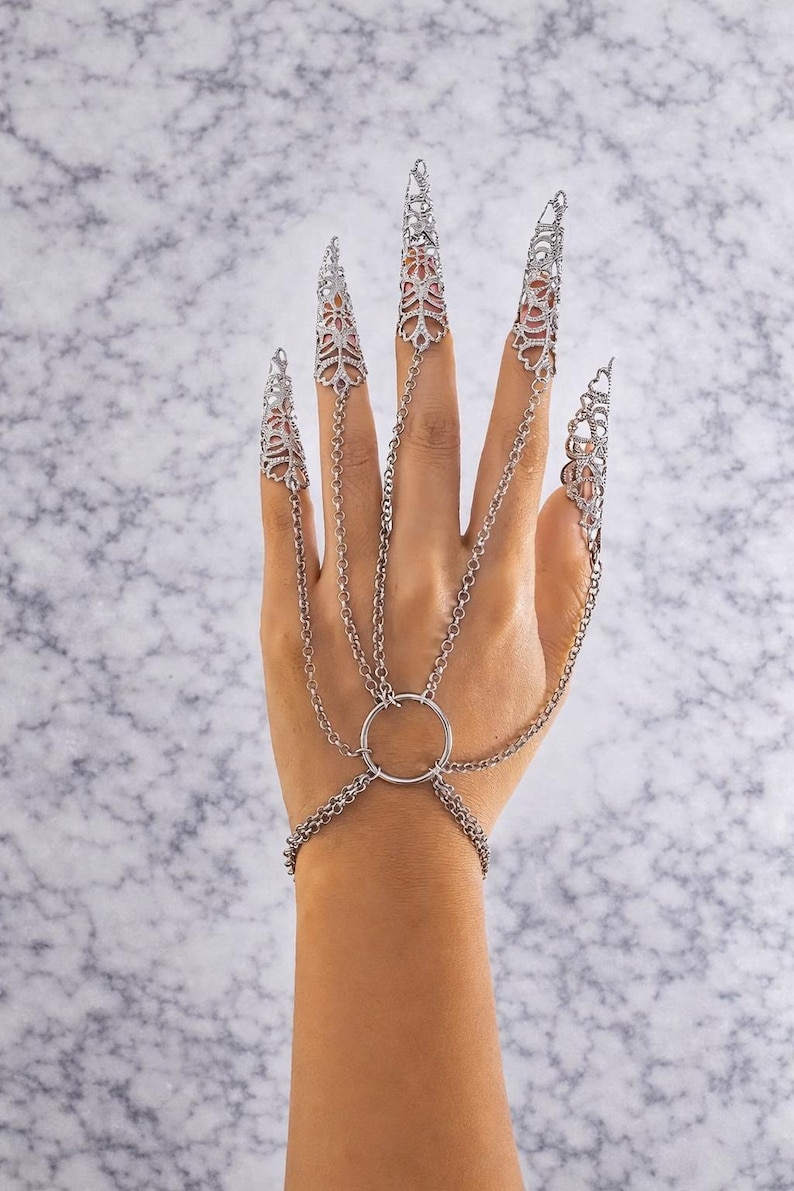 May include: Silver-toned finger armor jewelry with intricate filigree designs. The hand jewelry features chain links connecting the finger caps to a ring and bracelet, creating a unique and edgy accessory.