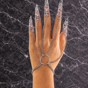 May include: Silver-toned hand jewelry featuring ornate, pointed finger covers connected by delicate chains to a wrist bracelet. The design includes a central ring and intricate filigree patterns, creating a gothic or fantasy aesthetic.