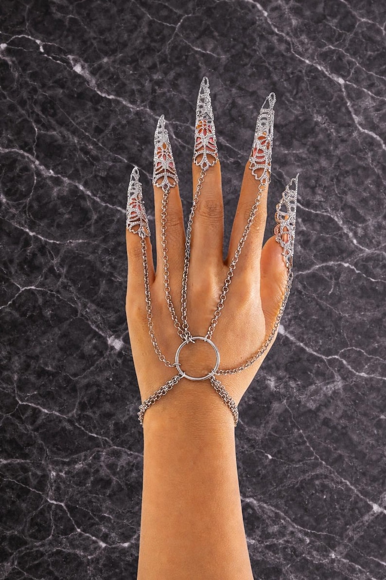May include: Silver-toned hand jewelry featuring ornate, pointed finger covers connected by delicate chains to a wrist bracelet. The design includes a central ring and intricate filigree patterns, creating a gothic or fantasy aesthetic.