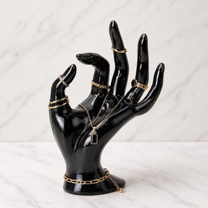 May include: A black, glossy hand-shaped jewelry display stand. The stand holds various gold rings, a necklace with a rectangular pendant, and a gold bracelet. The background is a white marble pattern.