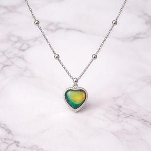 Peut inclure: Un collier argenté avec un pendentif en forme de cœur. Le pendentif change de couleur, passant du vert au jaune, et est entouré de petites pierres transparentes. La chaîne comporte de petites perles rondes.