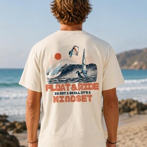 May include: Cream-colored t-shirt featuring a graphic of a wave, a sun, and windsurfers. The text "FLOAT & RIDE IS NOT A SKILL IT'S A MINDSET" is printed in orange. The shirt is worn by a person on a beach.