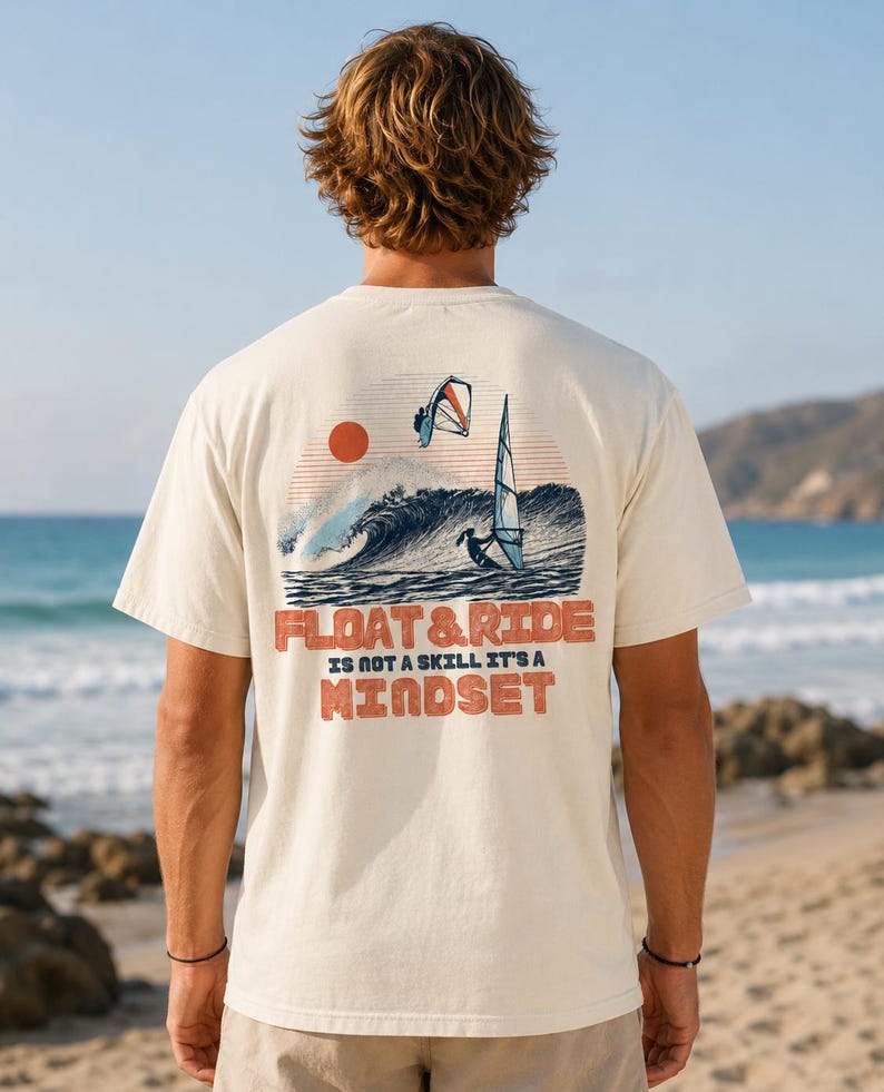 May include: Cream-colored t-shirt featuring a graphic of a wave, a sun, and windsurfers. The text "FLOAT & RIDE IS NOT A SKILL IT'S A MINDSET" is printed in orange. The shirt is worn by a person on a beach.