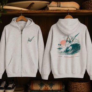May include: Light gray zip-up hoodie with a front pocket and a small teal and white graphic on the chest. The back features a larger graphic of a windsurfer on a wave with the text "WIND - WAVES - GOOD TIMES".