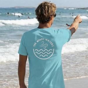 May include: A light blue t-shirt with the text "THE WINDY WATERS" in a white circular design. The design includes a wave and wind symbol. The shirt is worn by a person on a beach, with surfers in the background.