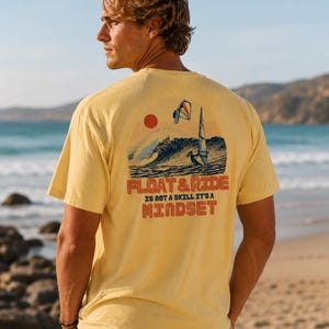May include: A yellow t-shirt with a graphic on the back featuring a sunset, ocean waves, and a windsurfer. The text "FLOAT & RIDE IS NOT A SKILL IT'S A MINDSET" is printed below the graphic. The shirt is worn by a person on a beach.