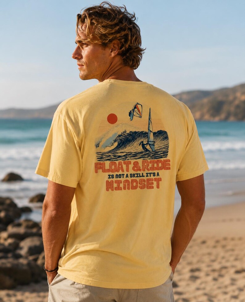 May include: A yellow t-shirt with a graphic on the back featuring a sunset, ocean waves, and a windsurfer. The text "FLOAT & RIDE IS NOT A SKILL IT'S A MINDSET" is printed below the graphic. The shirt is worn by a person on a beach.