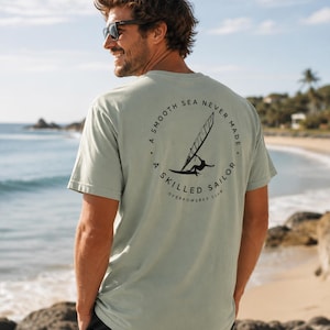 May include: A light green t-shirt with a circular graphic on the back featuring a windsurfer and the text "A SMOOTH SEA NEVER MADE A SKILLED SAILOR OVERPOWERED CLUB". The shirt is worn on a beach.