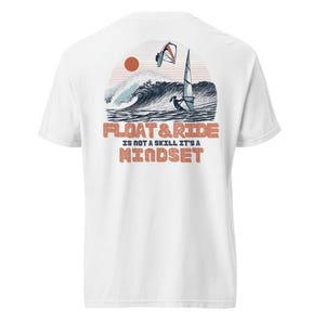 May include: White t-shirt with a graphic on the back featuring a windsurfer on a wave, a setting sun, and the text "FLOAT & RIDE IS NOT A SKILL IT'S A MINDSET" in orange and brown. The design evokes a beach and water sports theme.