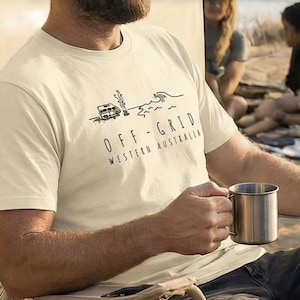 Może przedstawiać: Kremowa koszulka z czarną grafiką przedstawiającą kampera, fale i napis "OFF-GRID WESTERN AUSTRALIA". Osoba trzyma kubek ze stali nierdzewnej. Koszulka to element odzieży codziennej.