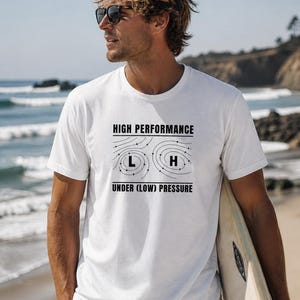 May include: White t-shirt with the text "HIGH PERFORMANCE UNDER (LOW) PRESSURE" in black. The shirt features a weather pattern graphic. The person is holding a surfboard on a beach.