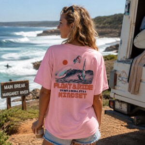 May include: A light pink t-shirt with a graphic on the back featuring a wave, sun, and windsurfer. The text "FLOAT & RIDE IS NOT A SKILL IT'S A MINDSET" is printed below the graphic. The shirt is worn outdoors near the ocean.