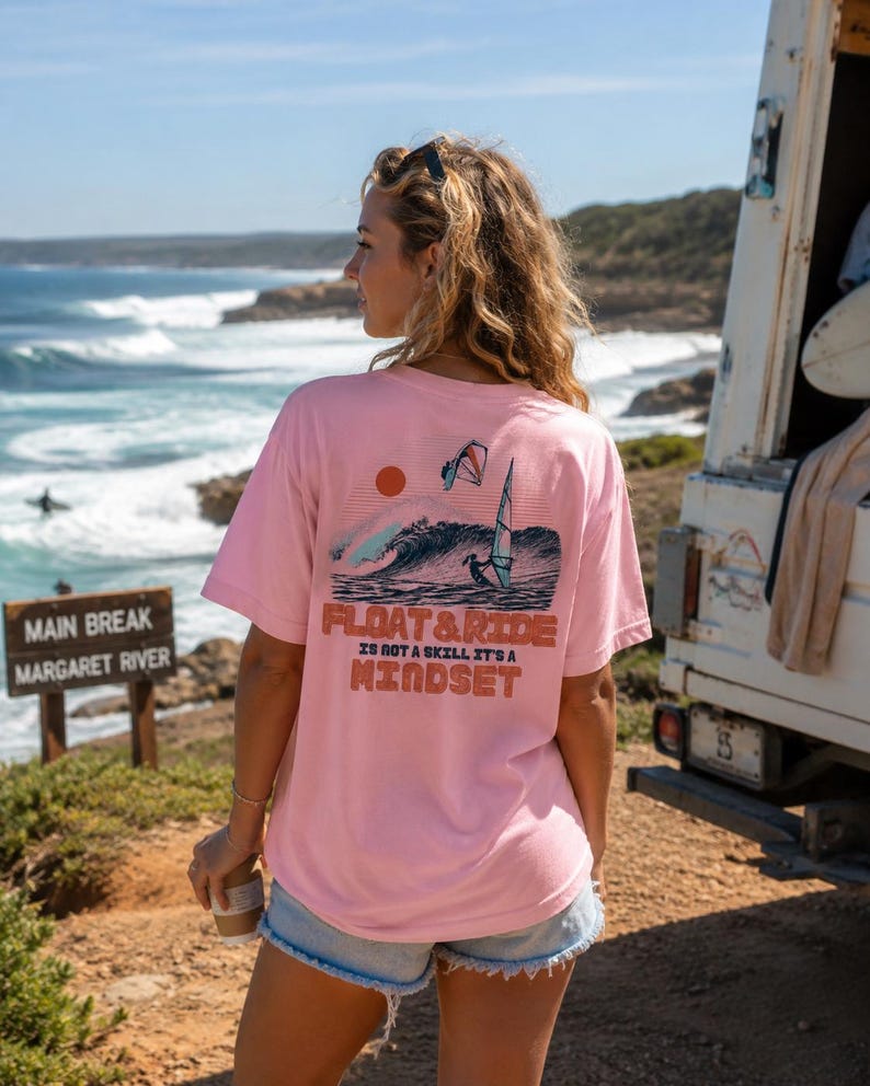 May include: A light pink t-shirt with a graphic on the back featuring a wave, sun, and windsurfer. The text "FLOAT & RIDE IS NOT A SKILL IT'S A MINDSET" is printed below the graphic. The shirt is worn outdoors near the ocean.