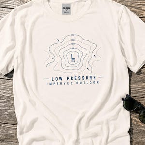May include: A white t-shirt with a navy blue graphic design. The design features a topographic map-like illustration with the letter "L" and the number "986" inside. Below the graphic, the text reads "LOW PRESSURE IMPROVES OUTLOOK".
