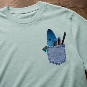 Op de afbeelding: Een lichtblauw t-shirt met een denim zak. De zak bevat een blauw surfboard, een kleine blauwe skateboard, twee zwarte voorwerpen en een paar zwart-grijze schoenen. Het shirt is gemaakt van een zacht materiaal.