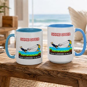 Può includere: Due tazze in ceramica bianca con manici e interni blu. Ogni tazza presenta una grafica "Game Over" con una scena di sport acquatici pixelata e il testo "Press Launch to Retry."