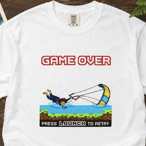 Può includere: T-shirt bianca con una grafica pixelata di una persona che cade in acqua mentre fa kitesurf. Le parole "GAME OVER" sono sopra la grafica in lettere pixelate rosse. Sotto la grafica, il testo recita "PRESS LAUNCH TO RETRY".