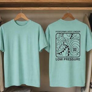 May include: Two light teal t-shirts hanging on wooden hangers. The front of one shirt is plain. The back of the other shirt has a black graphic with the text "PERFORMS BEST UNDER LOW PRESSURE".