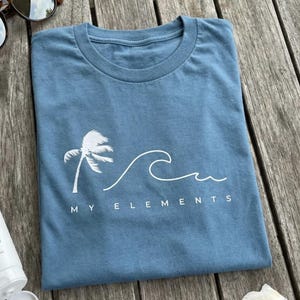 May include: A blue t-shirt with a white palm tree and wave design. The text "MY ELEMENTS" is printed below the wave. The shirt is folded and resting on a wooden surface.