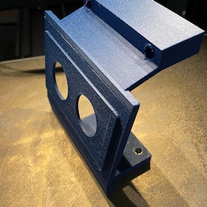Pode incluir: Um suporte azul escuro impresso em 3D com um painel vertical plano com dois recortes circulares e uma seção superior horizontal com dois pequenos orifícios circulares. O objeto apresenta uma textura em camadas típica da impressão 3D.