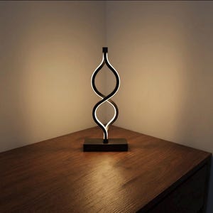 Peut inclure: Une lampe de table LED noire moderne avec un design unique et entrelacé. La lampe présente une base rectangulaire et un élément lumineux en forme de huit torsadé. La lumière émet une lueur douce et chaude, créant une pièce d'accent élégante. La lampe mesure environ 30 cm de haut.