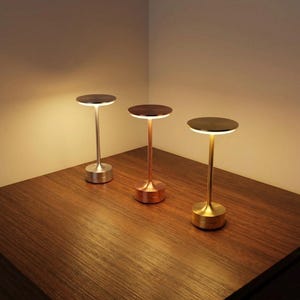 Peut inclure: Trois lampes de table modernes avec des abat-jours circulaires et des tiges fines. Les lampes ont des finitions métalliques argentées, cuivrées et dorées. Elles sont posées sur une surface en bois, éclairées par une lumière chaude.