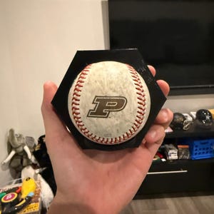 May include: A baseball with red stitching and a black "P" logo, set on a black hexagonal base. The baseball is off-white with visible wear. The base is held in a hand.