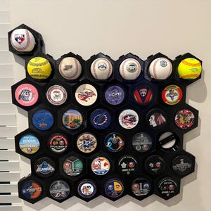 May include: A black, honeycomb-shaped display case mounted on a wall, showcasing baseballs, softballs, and hockey pucks. The display holds various sports memorabilia, including team logos and game-used items. The case is designed to hold multiple items.