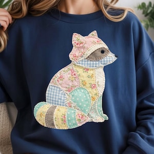 May include: Navy blue sweatshirt featuring a patchwork fox design. The fox is made of various patterned fabrics, including floral, polka dot, and gingham. The fox is sitting, with a light brown face and a striped tail. The sweatshirt is a solid navy blue color.