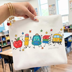 Peut inclure: Un t-shirt blanc avec un motif coloré représentant une pomme, un crayon, un sac à dos, une pile de livres et un bus scolaire souriants avec des bras et des jambes. Le design inclut des petites étoiles multicolores et le texte png.cology.
